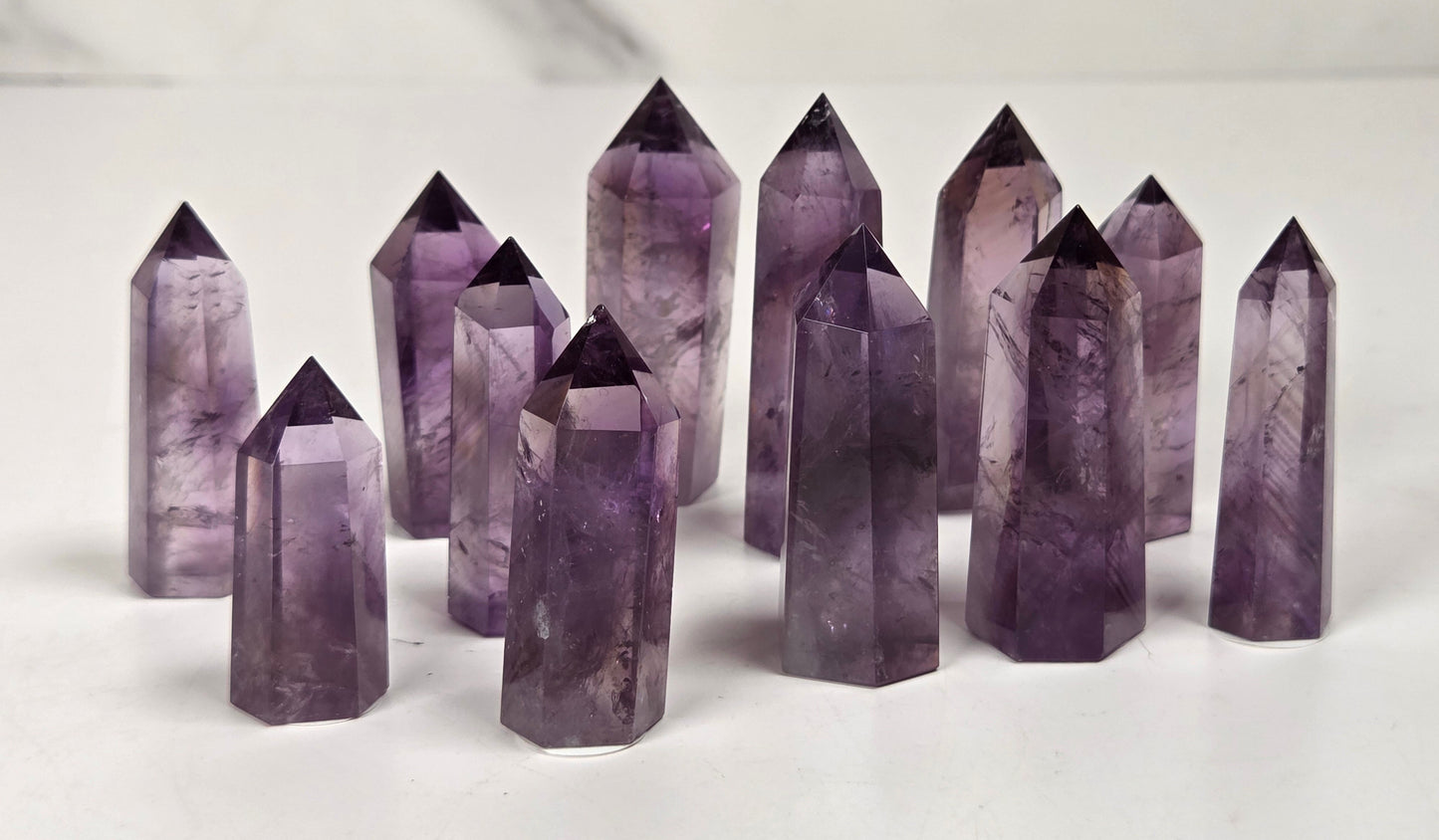 A group of natural amethyst crystal points of varying heights and sizes, showcasing translucent purple and lavender hues, arranged on a light marble surface.