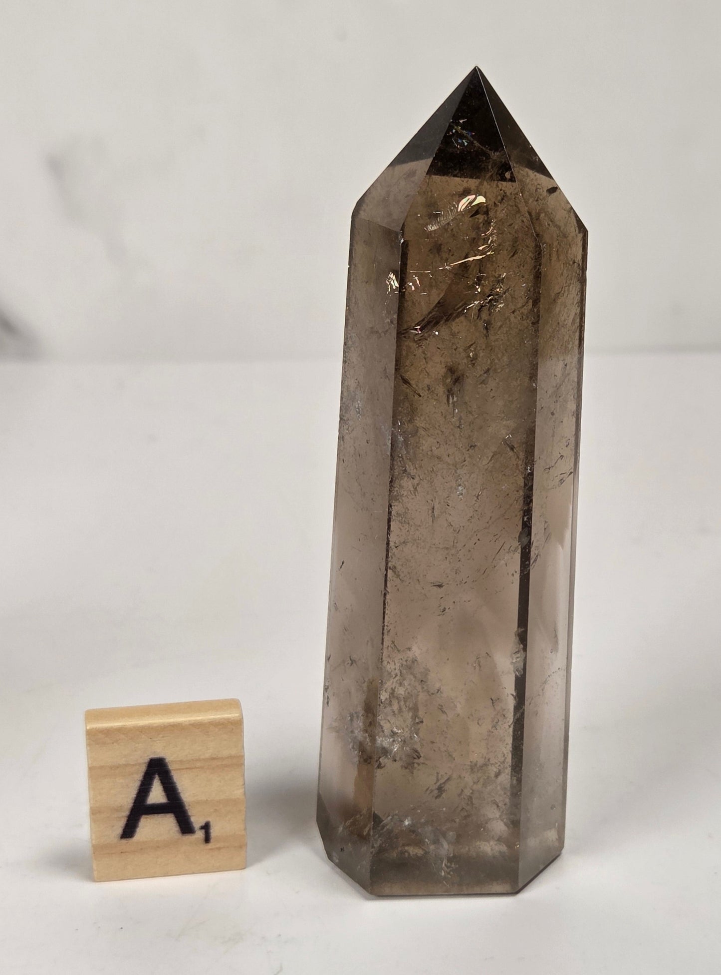 A tall, polished smoky quartz crystal point displayed on a light surface next to a small wooden letter tile with the letter 'A'.