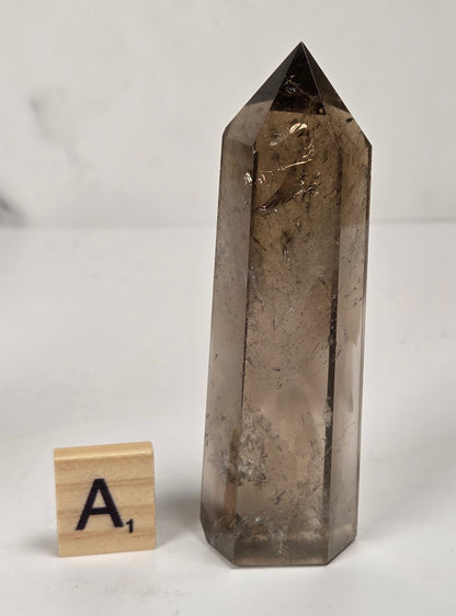 A tall, polished smoky quartz crystal point displayed on a light surface next to a small wooden letter tile with the letter 'A'.