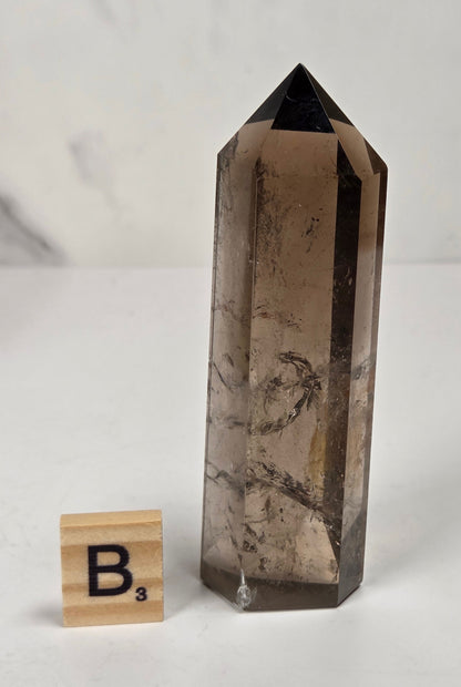 A smoky quartz crystal point tower with natural brown and black inclusions, shown on a white surface next to a small wooden Scrabble tile.