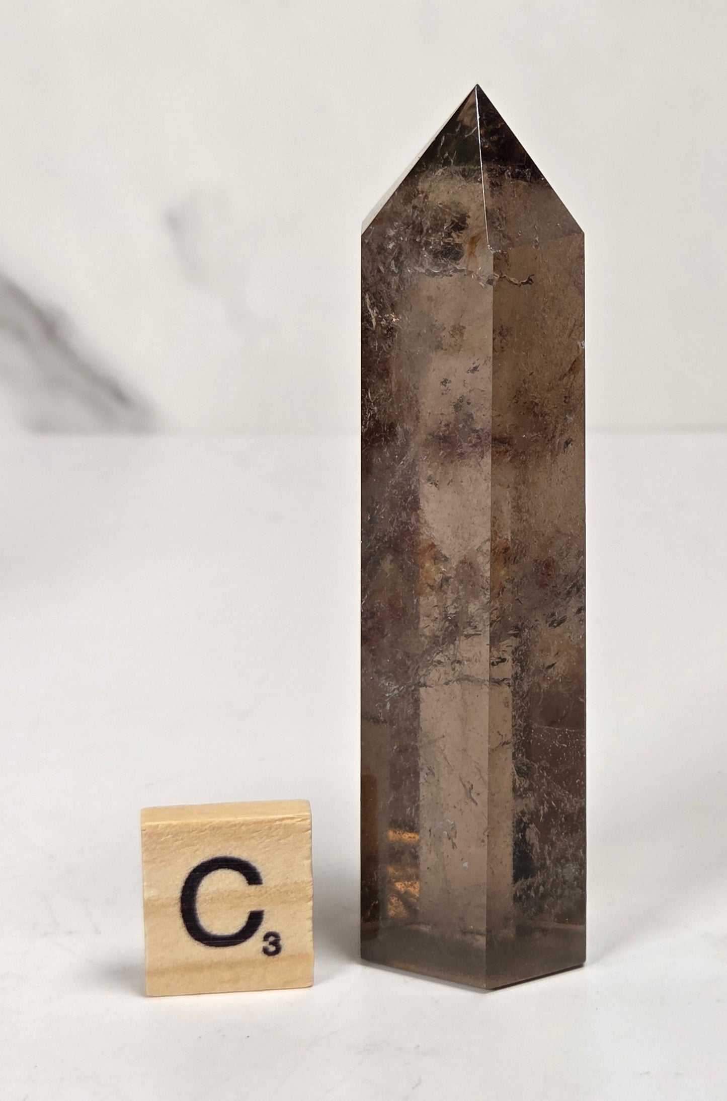 A smoky brown translucent crystal point tower standing upright on a white surface, with a small wooden Scrabble tile marked C³ beside it for scale, against a softly blurred light gray background.