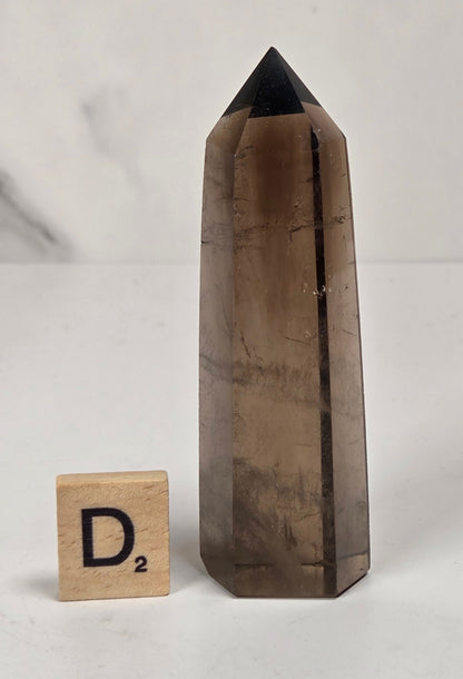 A tall, hexagonal smoky quartz crystal tower with a pointed black tip, shown next to a wooden Scrabble tile on a light marble surface.