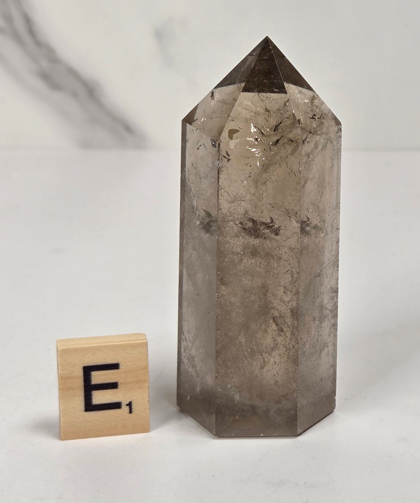 A natural smoky quartz crystal point with a faceted structure, shown next to a small wooden Scrabble tile on a light marble-patterned surface.