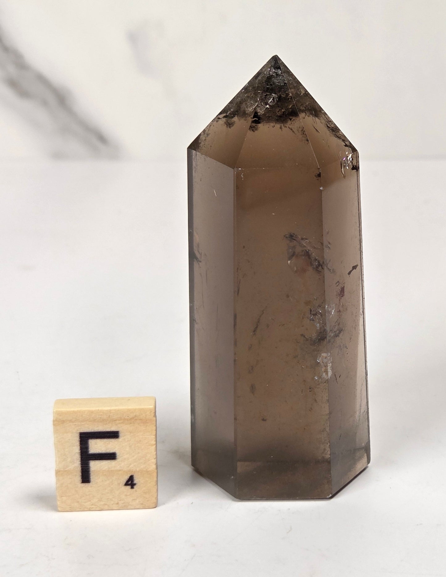 A smoky brown translucent crystal point tower with natural inclusions, shown next to a small wooden letter tile with the letter F on a light marble-like surface.