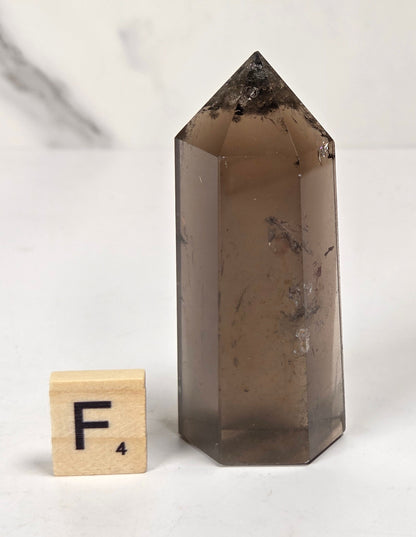 A smoky brown translucent crystal point tower with natural inclusions, shown next to a small wooden letter tile with the letter F on a light marble-like surface.