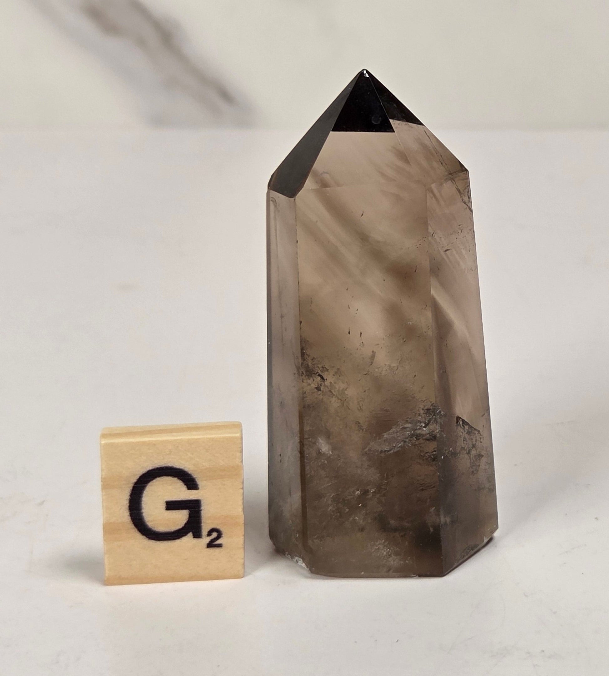 A smoky quartz crystal point with a triangular pyramid top, standing on a white surface next to a wooden Scrabble tile showing the letter 'G'.