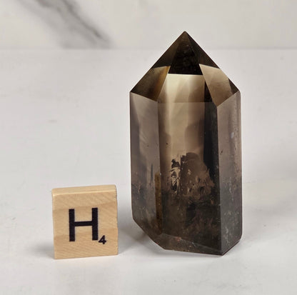 A smoky brown hexagonal crystal point with a natural landscape pattern inside, displayed next to a wooden Scrabble tile showing the letter 'H' on a light gray marble surface.