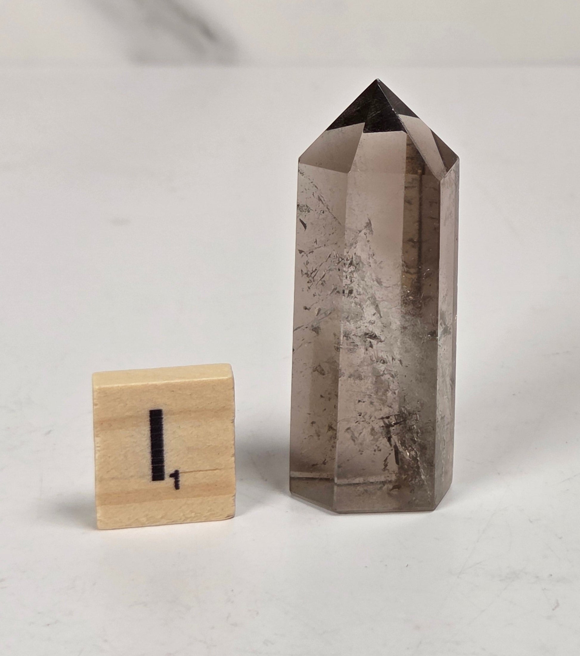 A tall, smoky quartz crystal point tower stands on a white surface next to a small wooden block with a black number one, set against a light gray marble background.