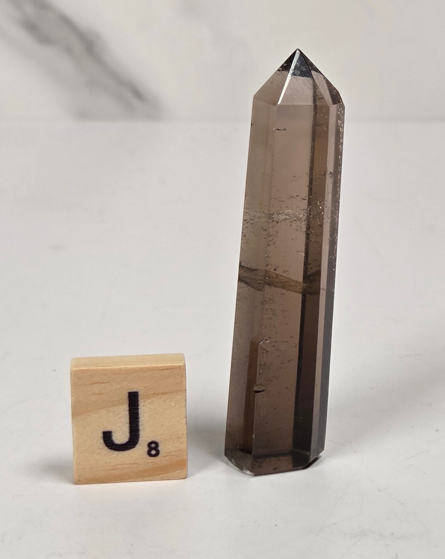 A smoky brown pointed crystal tower stands on a white surface next to a small wooden Scrabble tile with the letter J.