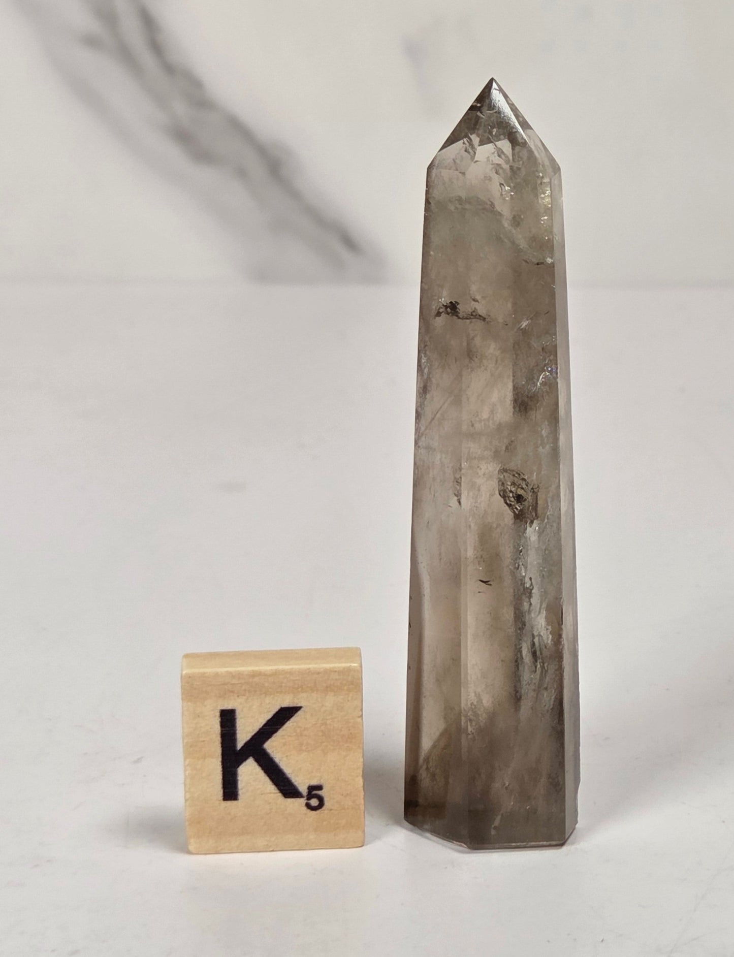 A tall, pointed smoky quartz crystal tower stands on a white surface next to a small wooden Scrabble tile showing the letter K.