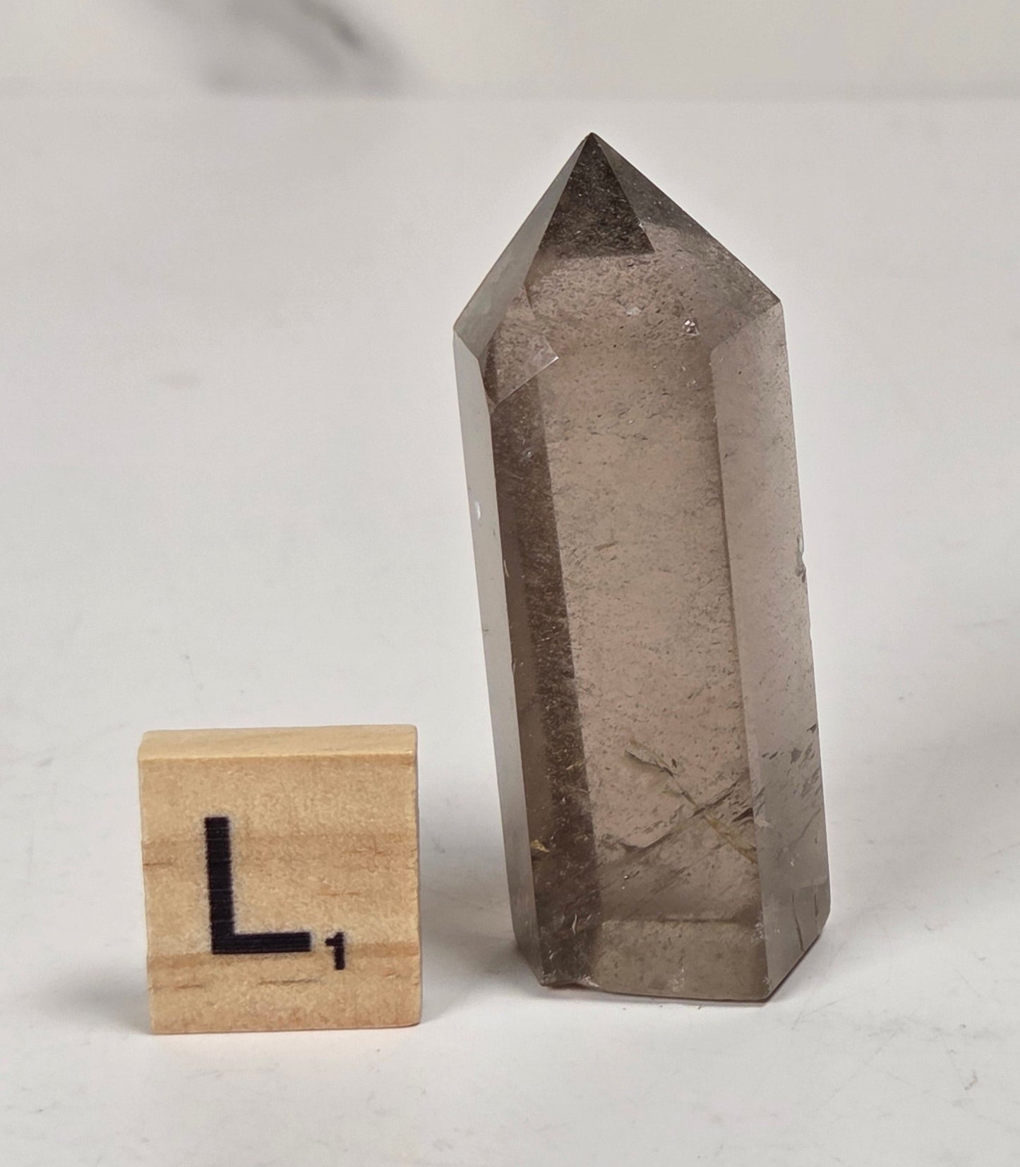 A smoky gray pointed crystal tower stands on a white surface next to a small wooden block with a black letter 'L' on it, against a light marble-patterned background.