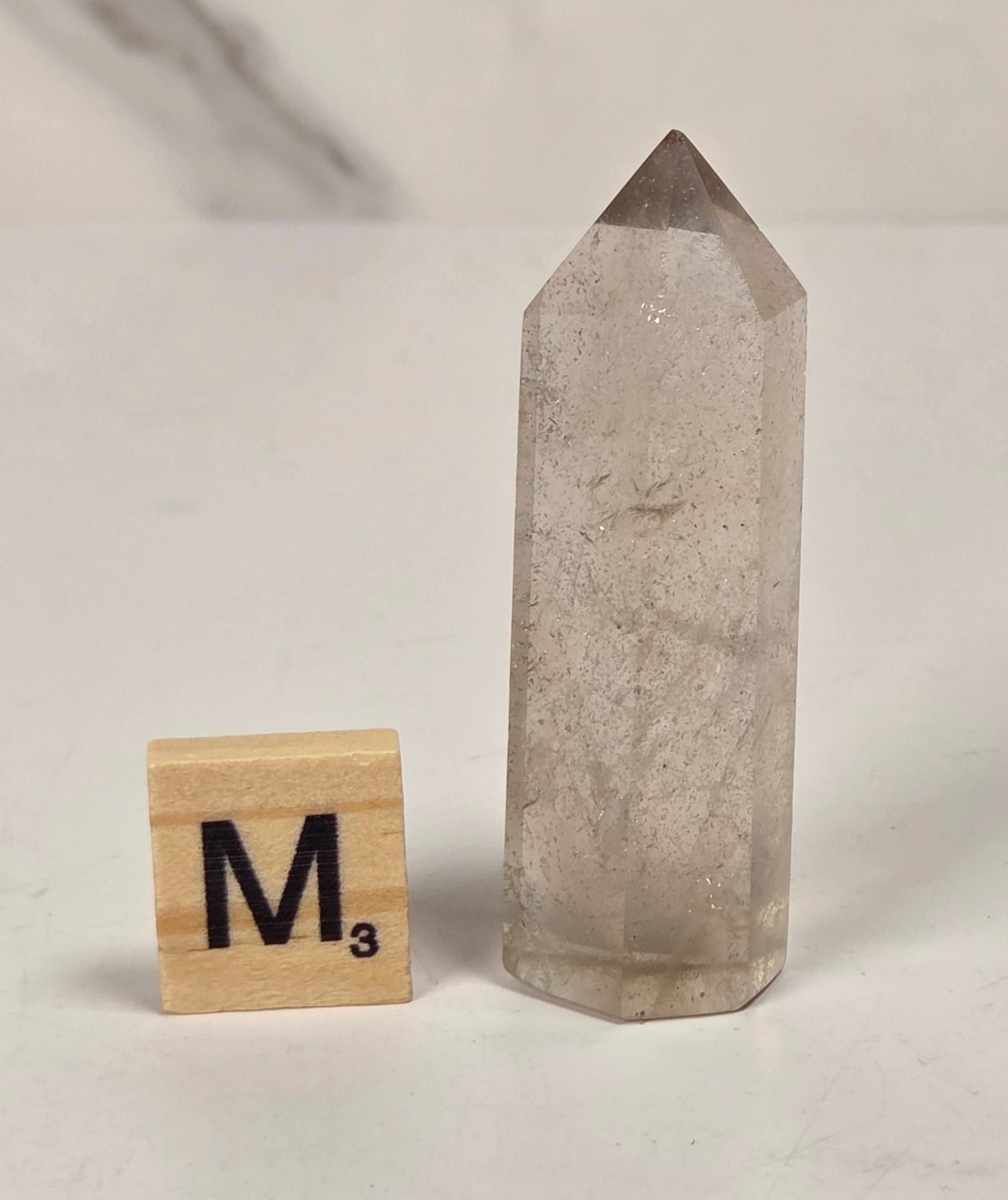A tall, pointed clear quartz crystal tower standing upright on a white surface, shown next to a small wooden Scrabble tile with the letter M. The background is a clean white surface with a soft, out-of-focus marble-like pattern.