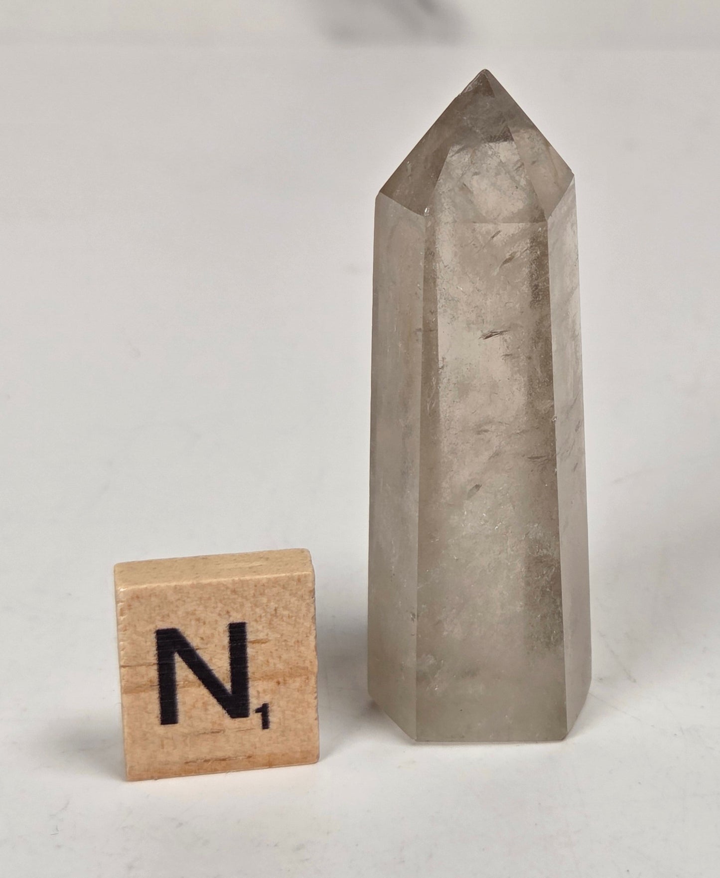 A tall, pointed clear quartz crystal tower standing upright on a white surface, with a small wooden Scrabble tile marked 'N1' placed beside it for scale.