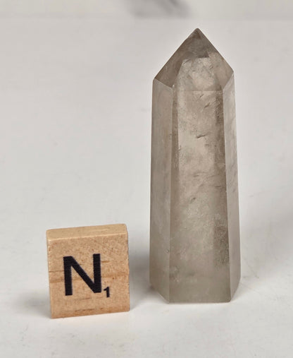 A tall, pointed clear quartz crystal tower standing upright on a white surface, with a small wooden Scrabble tile marked 'N1' placed beside it for scale.
