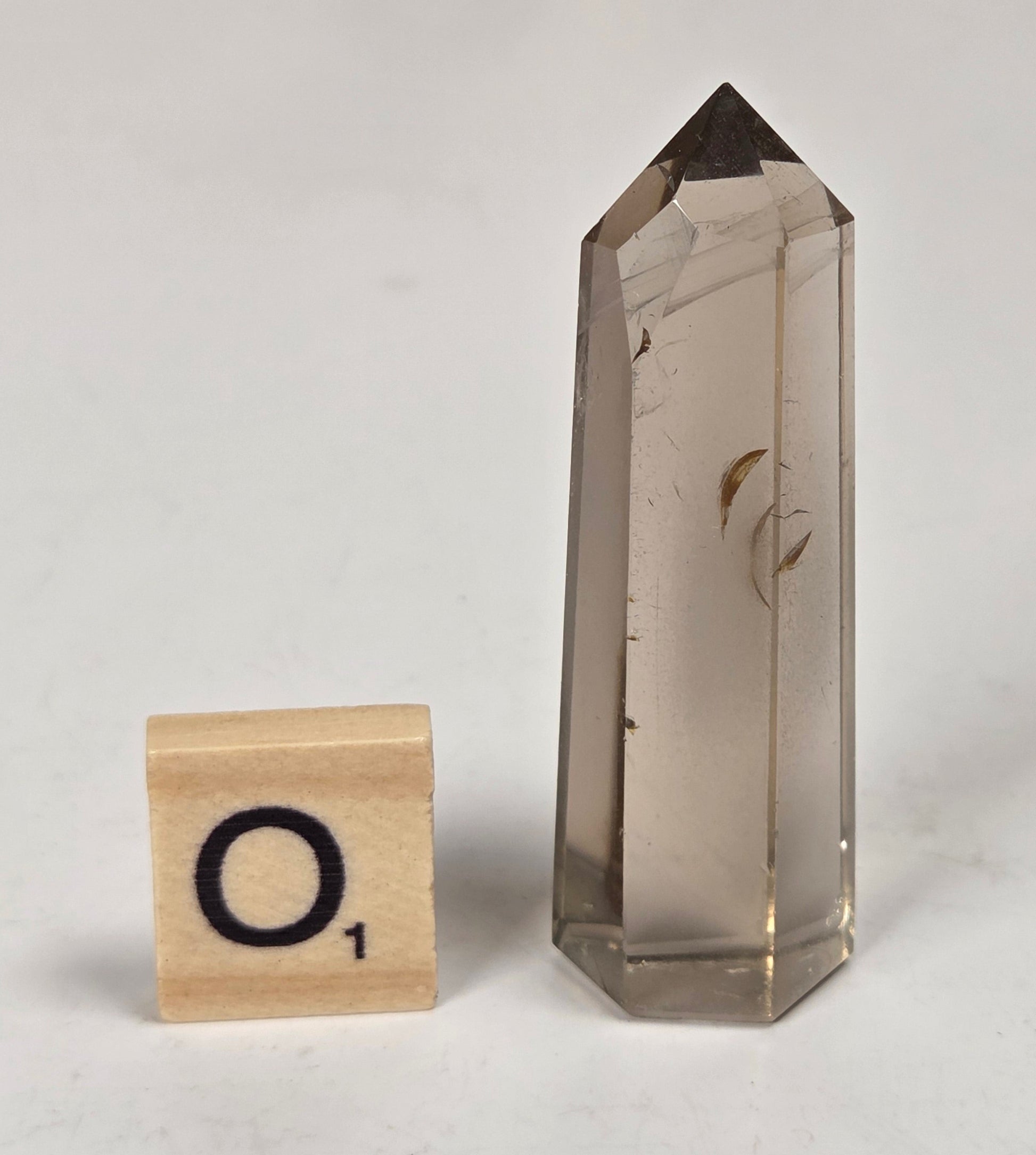 A smoky quartz crystal point with natural inclusions, shown next to a wooden Scrabble tile with the letter 'O' on a light gray marble-like surface.