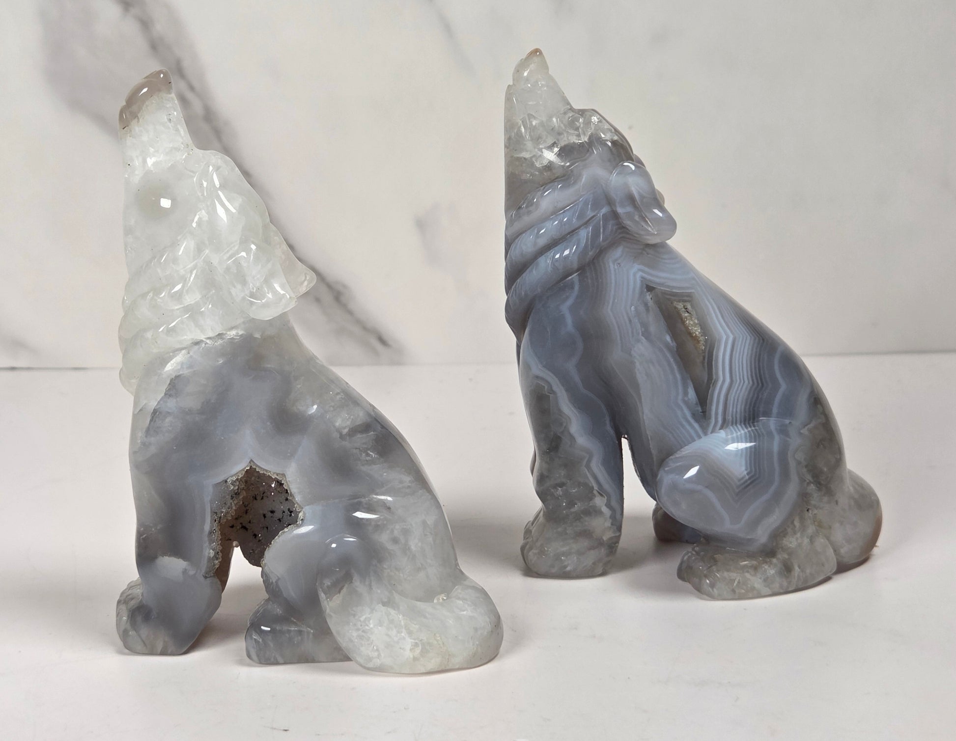 Two carved stone animal figurines, shaped like seated foxes or hounds, displayed on a white surface against a light marble-patterned background.
