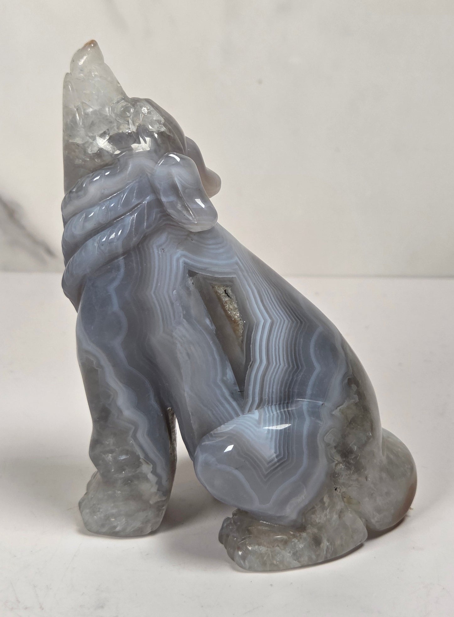 A small carved stone figurine of a seated animal, likely a fox or wolf, made from layered gray agate with natural banding and a clear crystal pointed upward, shown on a light surface against a marble-look background.