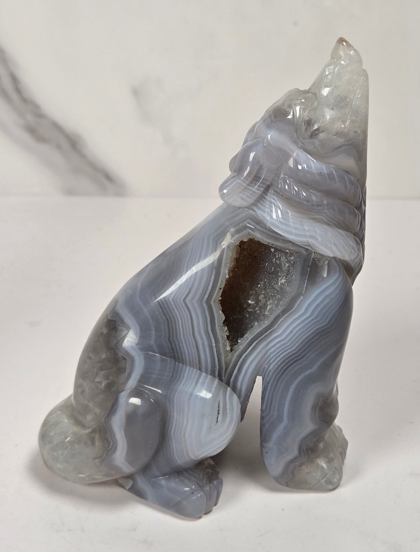 A carved stone figurine of a seated animal, likely a fox or wolf, made from polished gray agate with natural banded striations and a rough crystalline cavity opening, shown on a light marble surface.