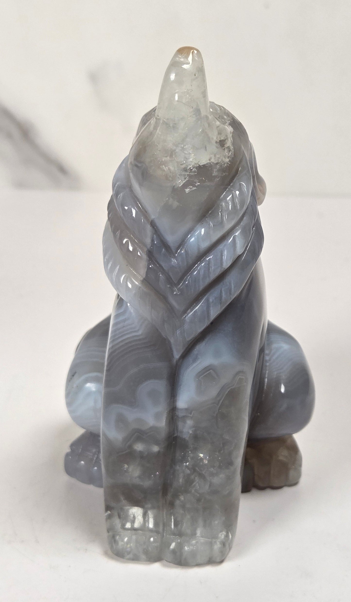 A small carved crystal figurine of a wolf or fox in a crouched position, made from marbled gray agate with a clear quartz crystal point on its head, set against a light marble background.