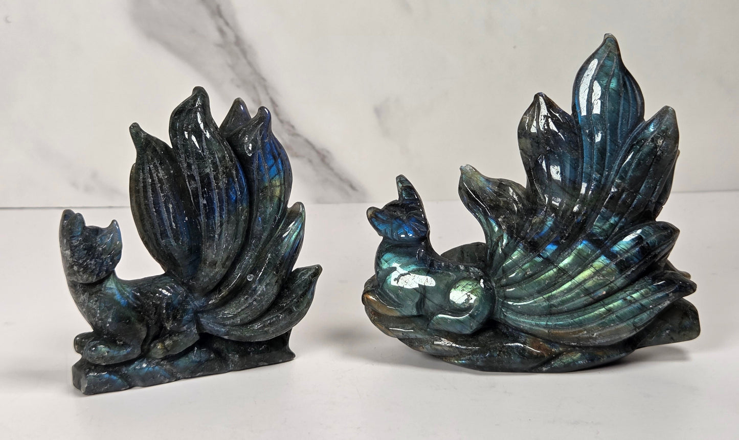 Two Labradorite figurines shaped like a fox and a peacock, displayed side by side on a white surface with a white and gray marble background.