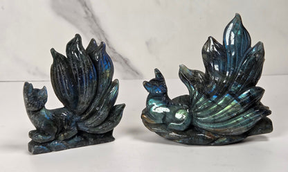 Two Labradorite figurines shaped like a fox and a peacock, displayed side by side on a white surface with a white and gray marble background.