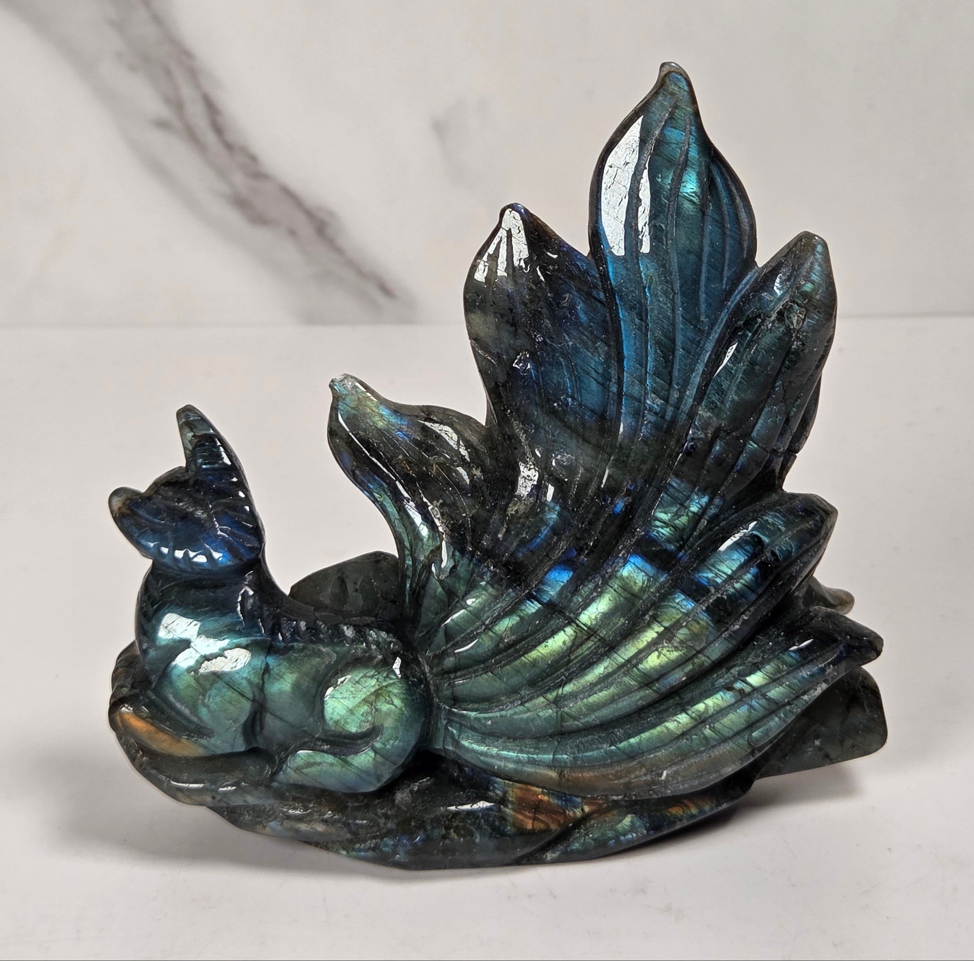 A polished labradorite figurine shaped like a small creature with a large, fan-like back, displayed on a white marble surface with a white marble background.