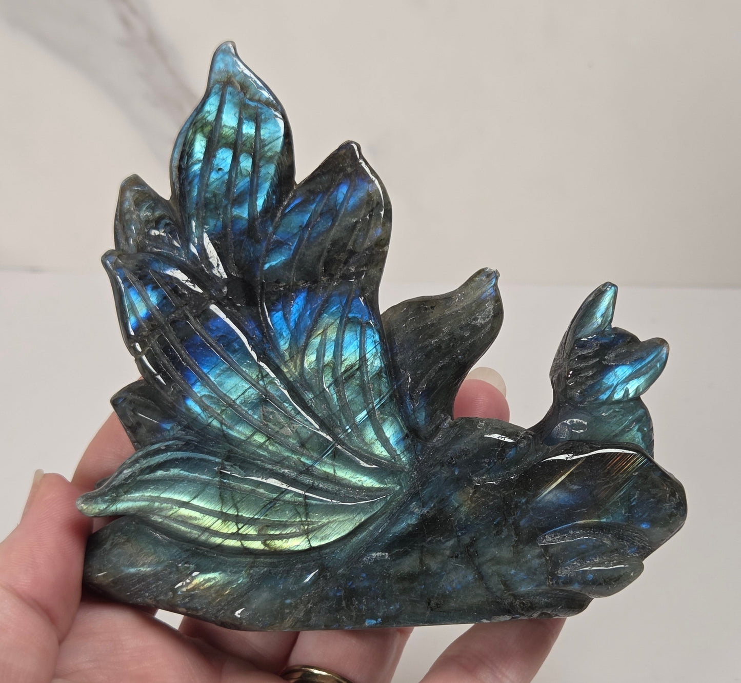 A hand holding a polished labradorite crystal cluster shaped like leaves, showing a shimmering blue-green iridescent finish against a light neutral background.