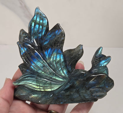 A hand holding a polished labradorite crystal cluster shaped like leaves, showing a shimmering blue-green iridescent finish against a light neutral background.