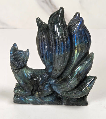 A small carved figurine of a fox with a large bush of leaves or petals behind it, made from iridescent labradorite stone showing blue, green, and purple flashes, set against a white marble-patterned background.