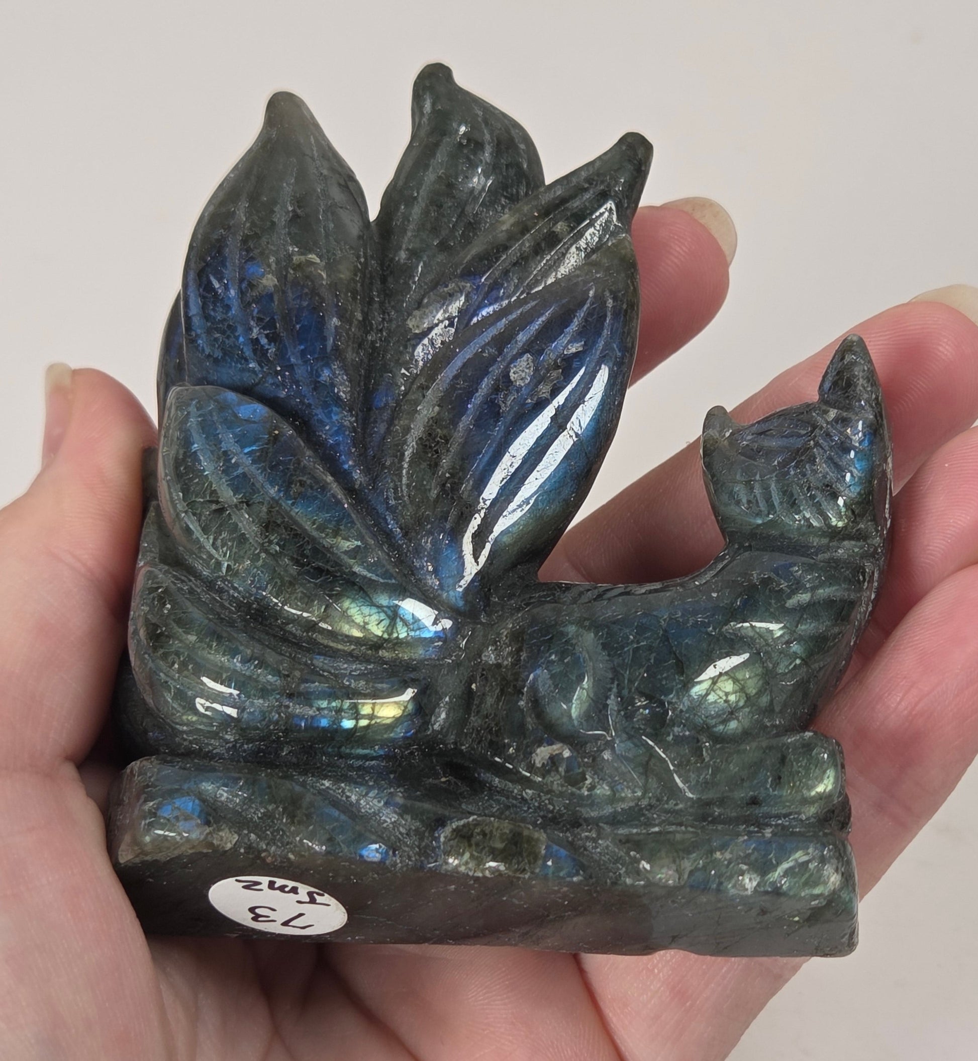 A hand holding a small carved labradorite figurine shaped like a fox with a lotus flower above it. The stone shows an iridescent blue, green, and gray play-of-color effect, with a small white price sticker on the base.