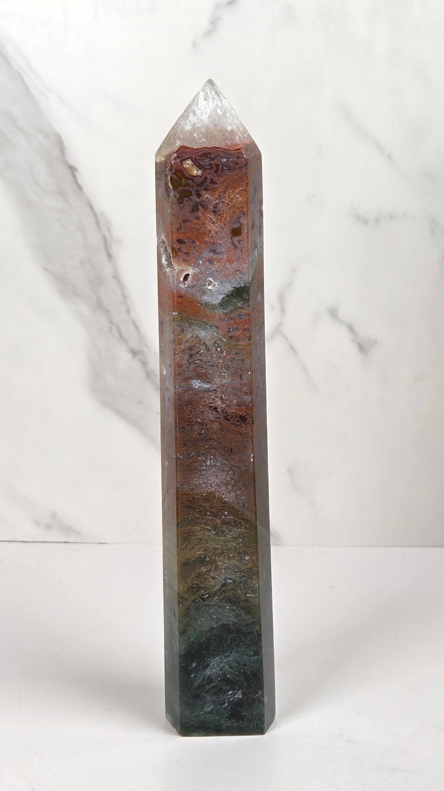 A tall, polished crystal tower with a pointed tip, featuring natural earthy coloration in shades of brown, gray, and dark green, standing on a white surface against a light gray marble-patterned background.
