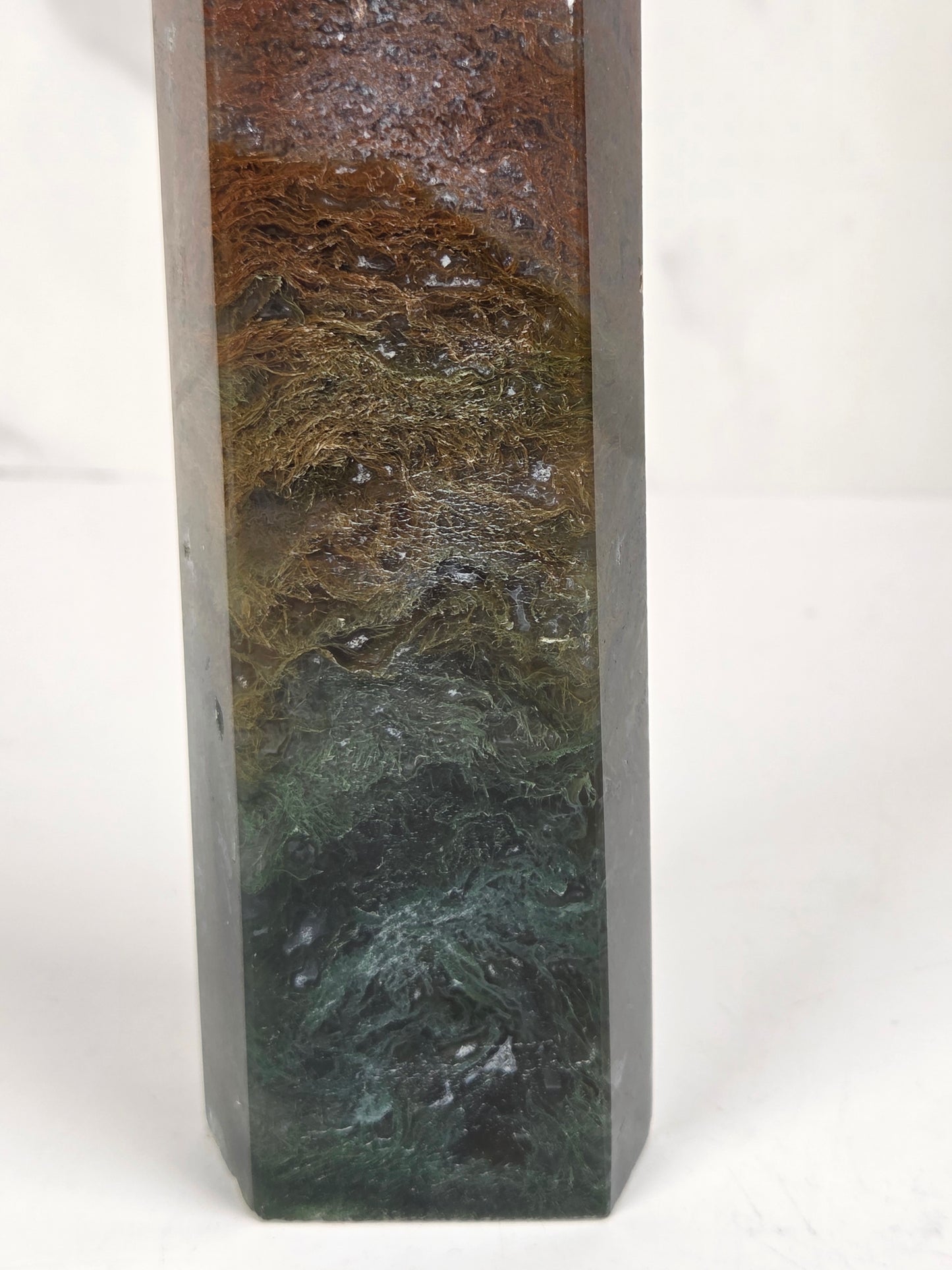close-up of a tall, polished crystal tower with a pointed tip, featuring natural earthy coloration in shades of brown, gray, and dark green, standing on a white surface against a light gray marble-patterned background.