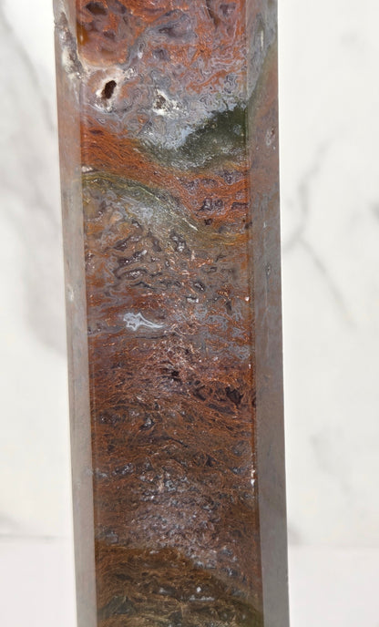 A close-up of a polished rectangular stone column featuring natural marbled patterns in earthy brown, rust orange, gray, and olive green, set against a light marble-patterned background.