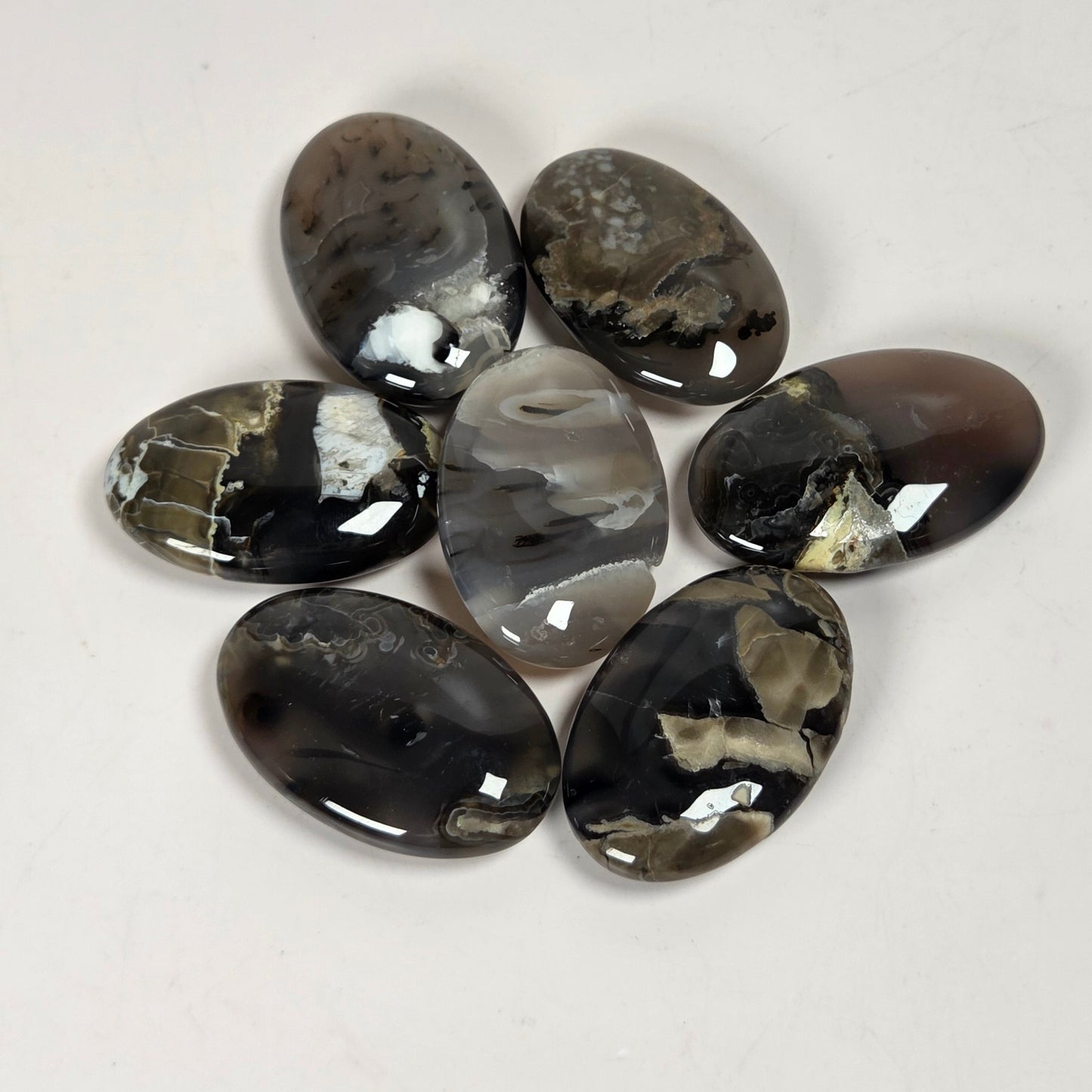 Seven polished oval agate stones with natural marbled patterns in black, gray, white, and brown, arranged in a circle on a plain off-white background.