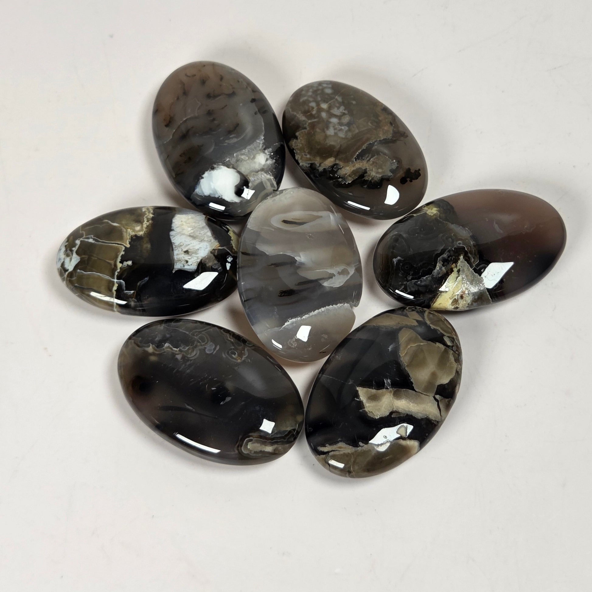 Seven polished oval agate stones with natural marbled patterns in black, gray, white, and brown, arranged in a circle on a plain off-white background.