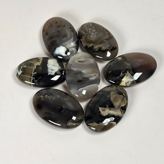 Seven polished oval agate stones with natural marbled patterns in black, gray, white, and brown, arranged in a circle on a plain off-white background.