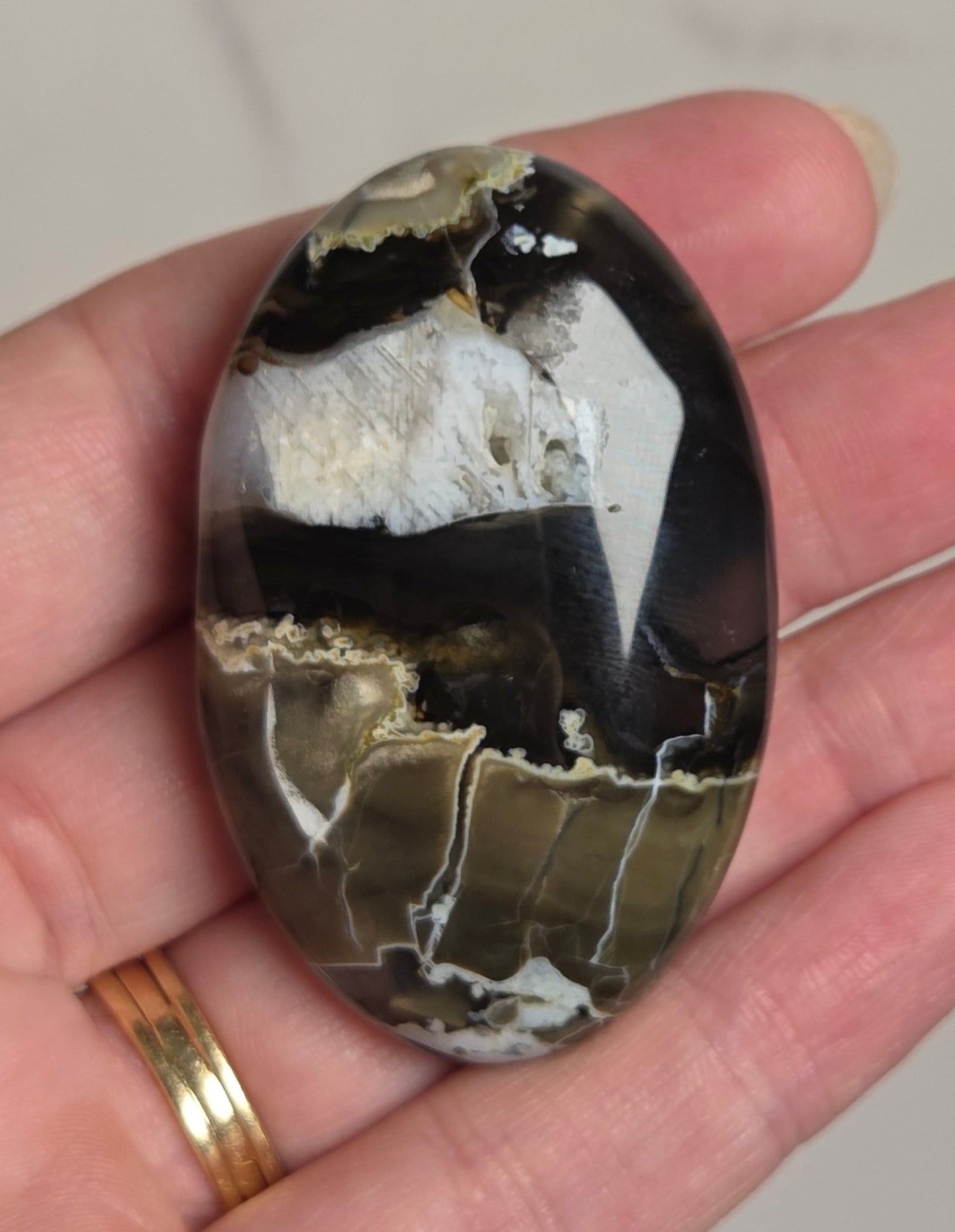 A polished oval stone with black, white, and olive-brown banding patterns, held in a person's hand against a light marble background.