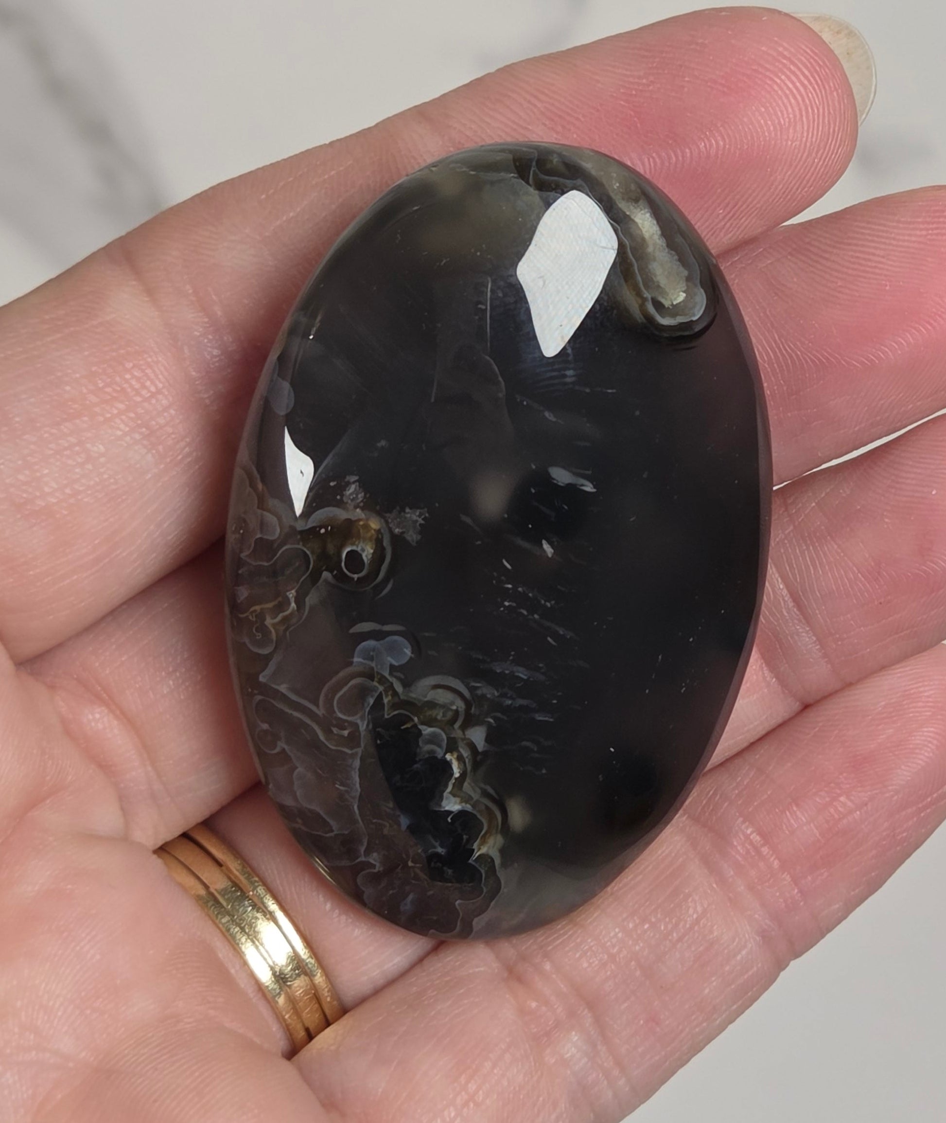 A hand holding a polished oval cabochon gemstone with a dark, marbled pattern of black, gray, and brown, set against a light marble background.