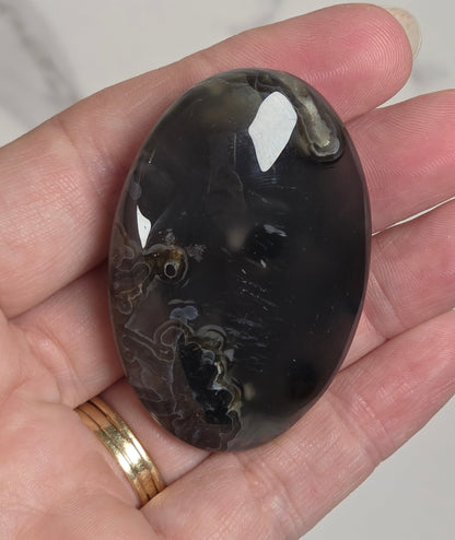 A hand holding a polished oval cabochon gemstone with a dark, marbled pattern of black, gray, and brown, set against a light marble background.