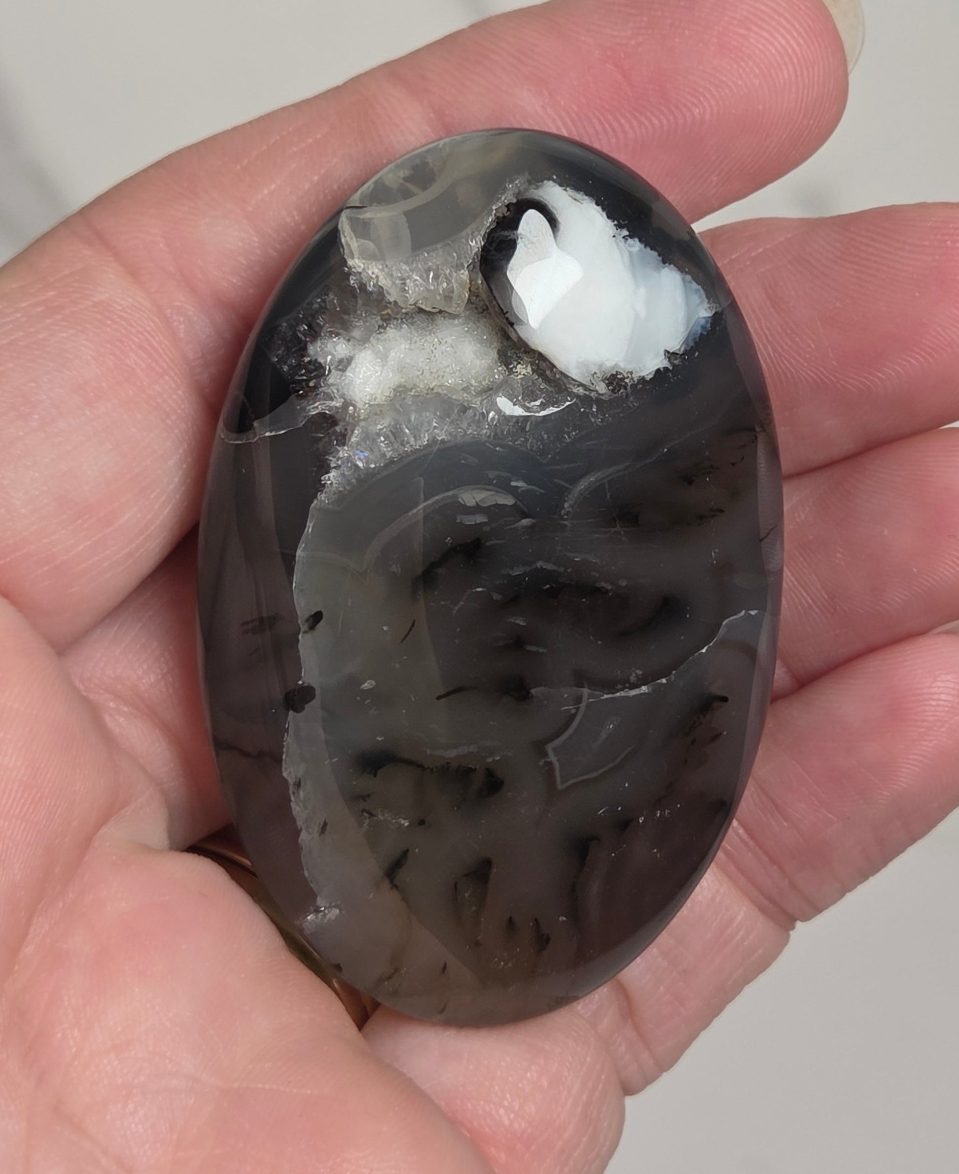 A hand holding a polished oval agate stone with natural marbled patterns in black, gray, and white, set against a light marble-patterned background.