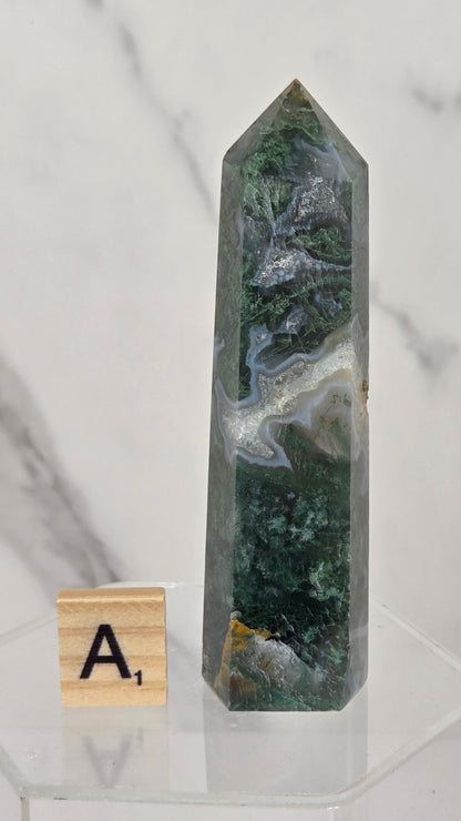 A polished natural crystal tower with green, gray, and white agate-like banding stands on a clear acrylic display base next to a small wooden letter tile showing 'A1'. The background is a white marble surface and wall.