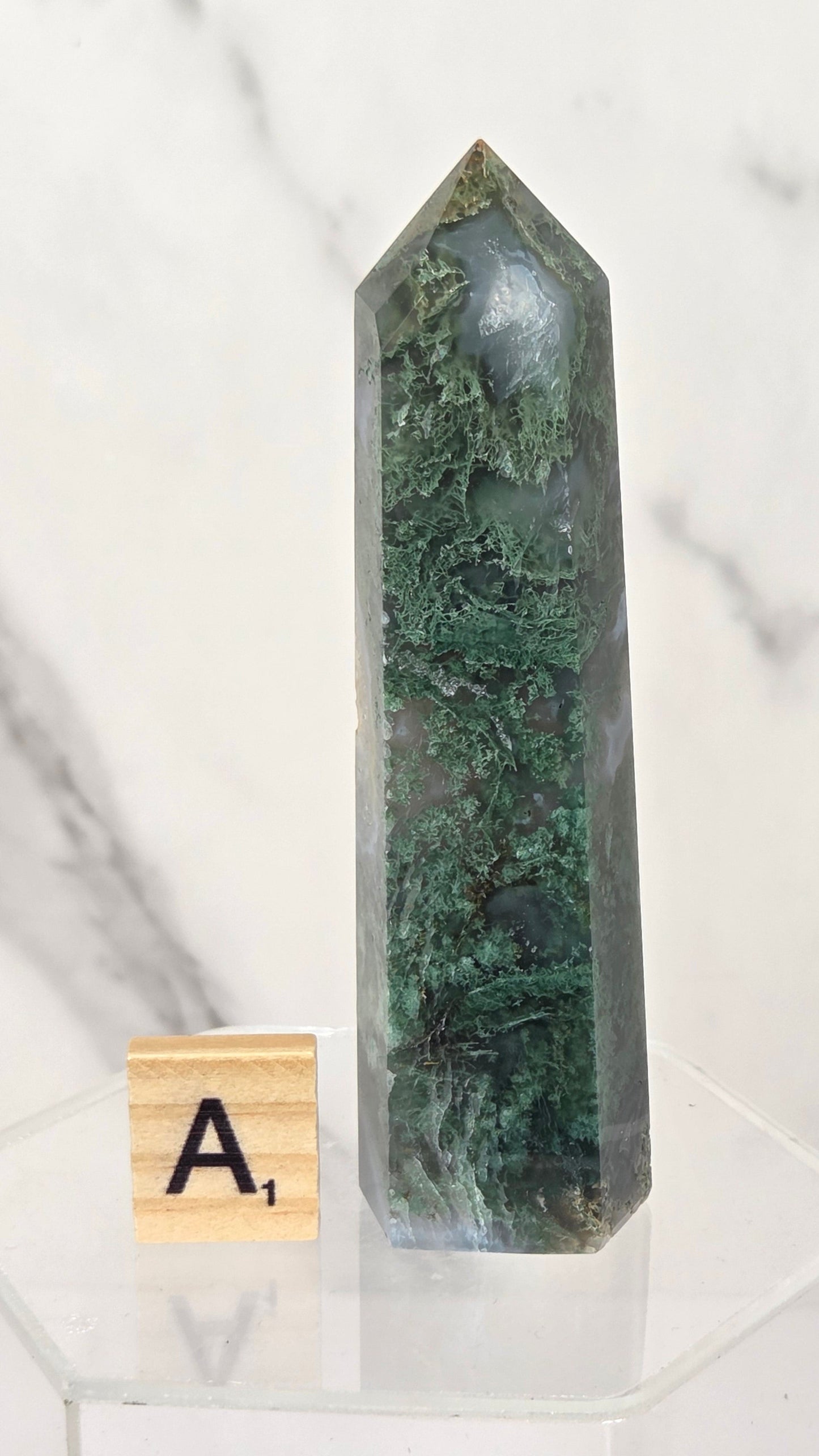 A green moss agate crystal point tower displayed on a clear acrylic stand next to a wooden Scrabble tile showing the letter 'A', set against a white and gray marble background.
