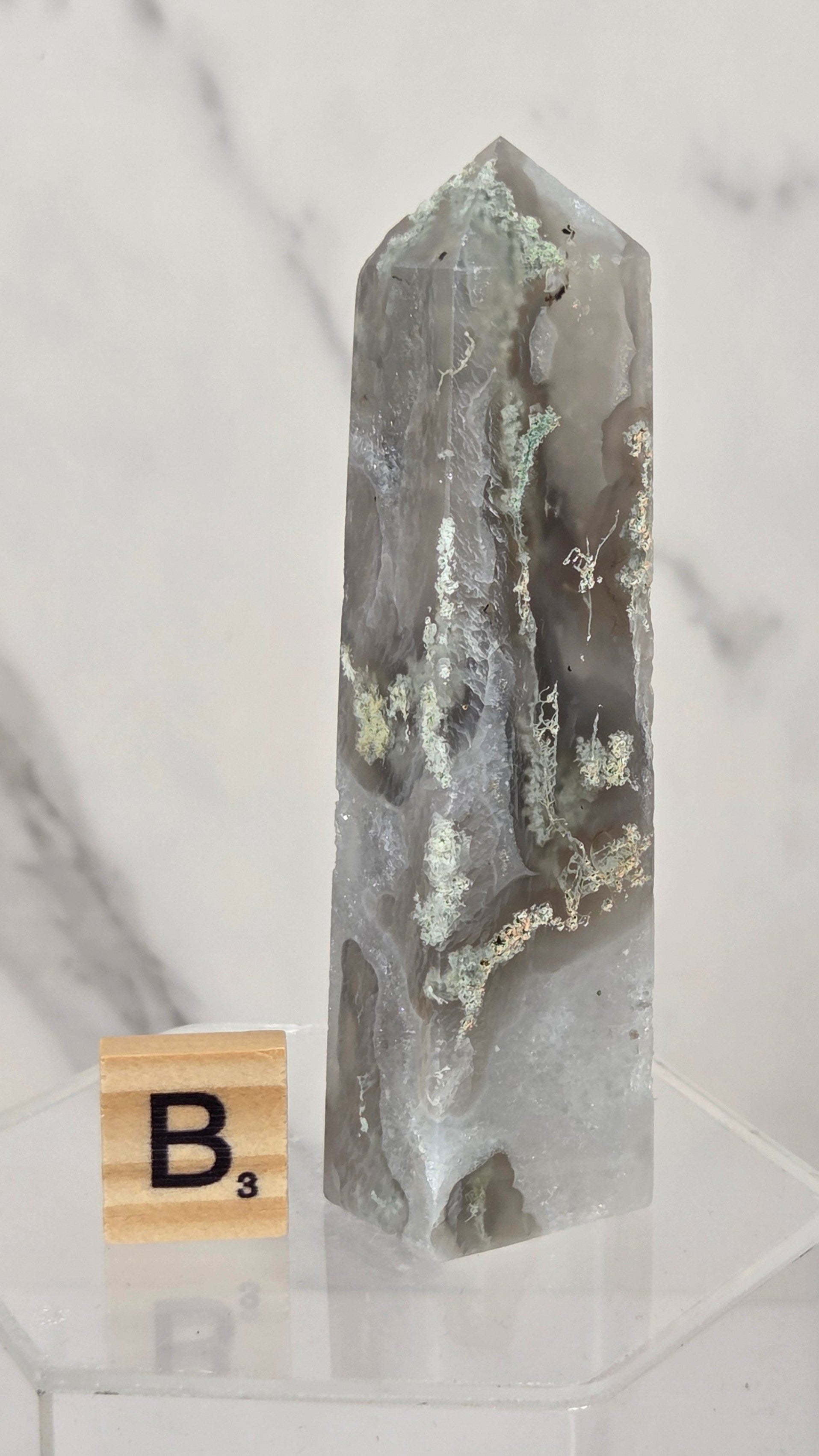 A tall, pointed gray agate crystal tower with natural inclusions and a metallic silver point, displayed on a clear acrylic stand next to a wooden Scrabble tile.