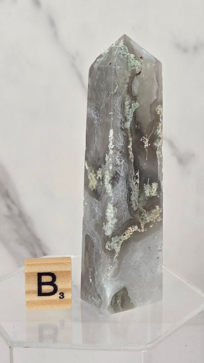 A tall, pointed gray agate crystal tower with natural inclusions and a metallic silver point, displayed on a clear acrylic stand next to a wooden Scrabble tile.