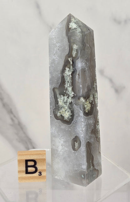 A tall, pointed clear quartz crystal tower with natural gray and white agate-like inclusions, displayed on a clear acrylic stand next to a wooden Scrabble tile marked B3, set against a light gray marble background.