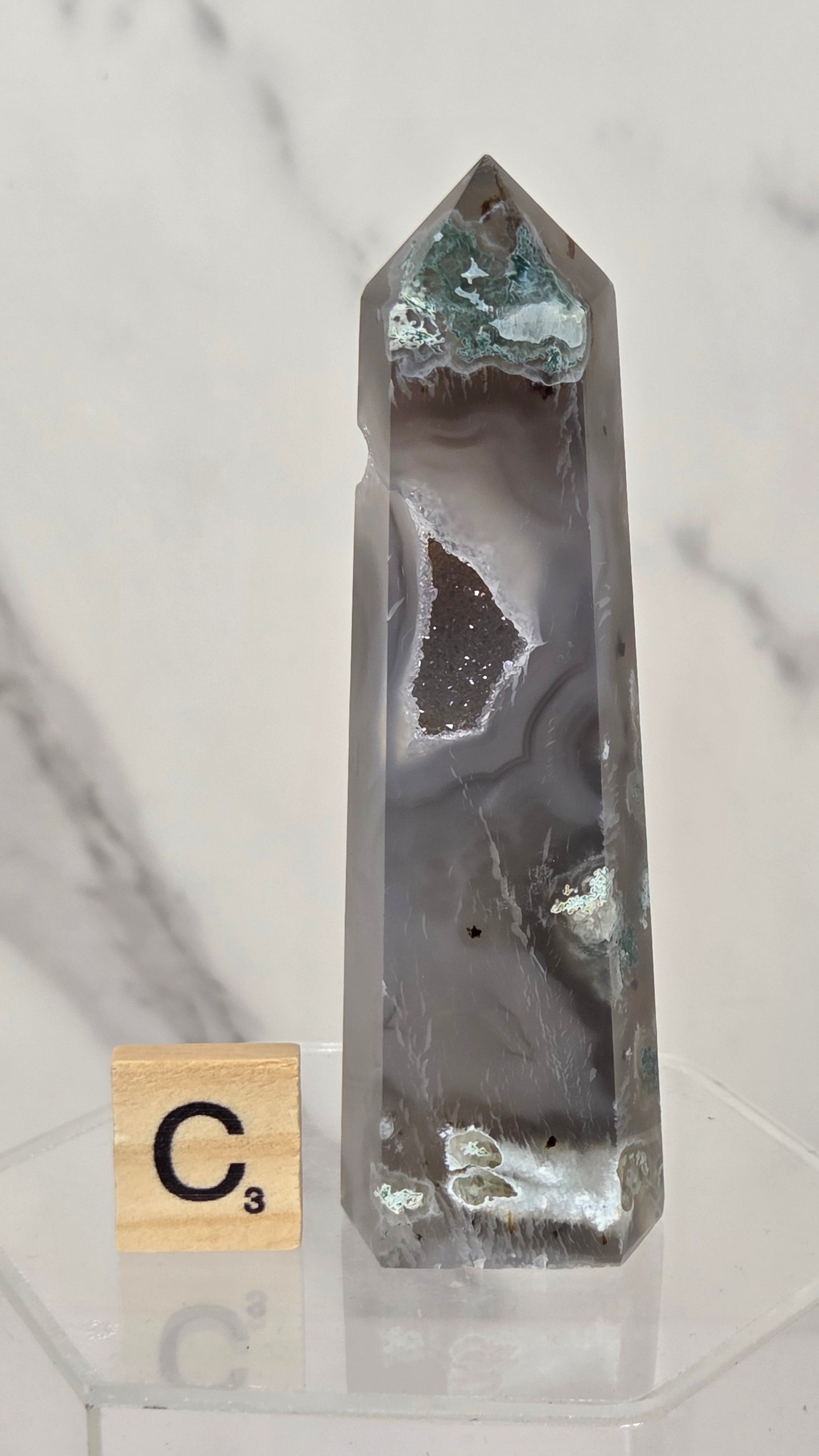 A tall, pointed clear quartz crystal point with natural agate banding and metallic silver inclusions, displayed on a clear acrylic stand next to a Scrabble tile showing the letter C for cost.