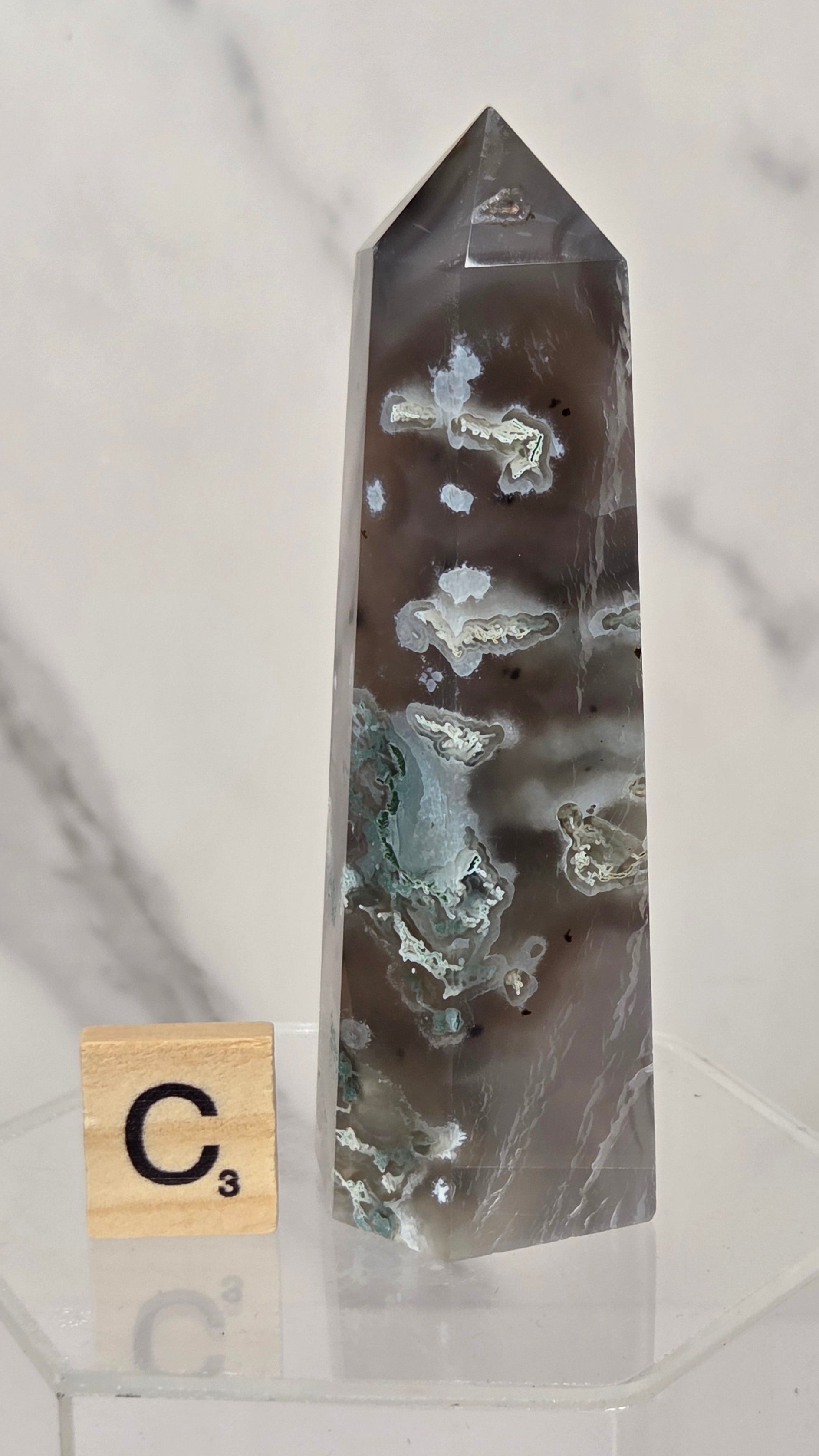 A tall, pointed clear crystal tower with natural white and gray mineral inclusions, displayed on a clear acrylic stand next to a small Scrabble tile showing the letter C3, against a white marble background.