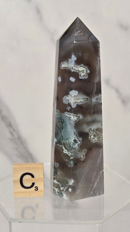 A tall, pointed clear crystal tower with natural white and gray mineral inclusions, displayed on a clear acrylic stand next to a small Scrabble tile showing the letter C3, against a white marble background.