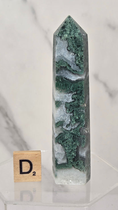A polished green and white marbled crystal point tower displayed on a clear acrylic stand next to a small wooden Scrabble tile showing the letter D2, set against a white marble background.