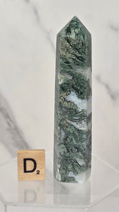 A tall, pointed green crystal tower with natural marbled patterns, displayed on a clear acrylic stand next to a wooden Scrabble tile showing the letter 'D2', set against a white marble background.